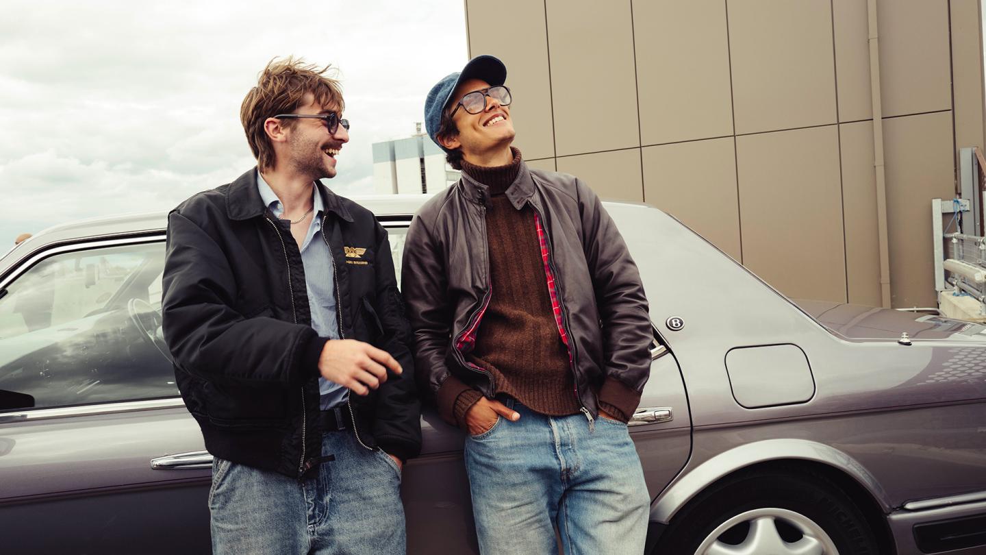 Two men smiling and leaning against a vintage car on a cloudy day