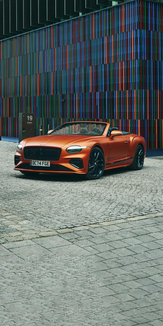 An orange Continental GTC is parked in front of a colourful modern building